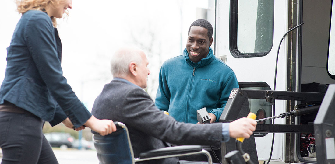 A smiling Impark HEALTH attendant assisting a man in a wheelchair who is boarding an Impark HEALTH shuttle bus.
